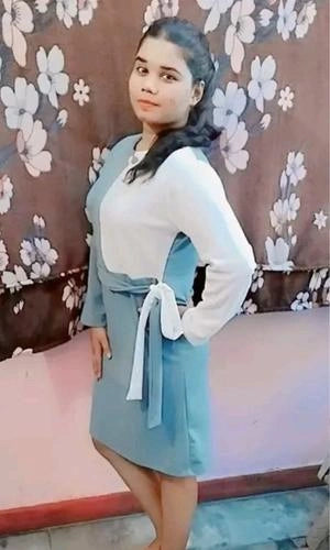 Woman wearing a light blue dress with a white long-sleeve shirt underneath, standing against a floral-patterned wall.