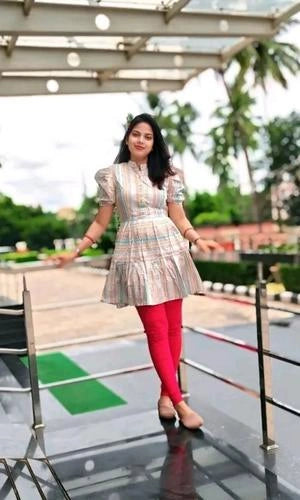 Woman in a patterned dress and pink leggings standing on a reflective surface with greenery in the background