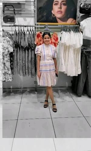 Woman in a patterned dress standing in a clothing store with racks of clothes and a poster on the wall.