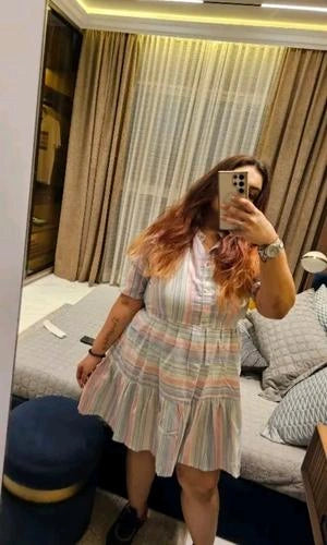 Person wearing a striped dress taking a mirror selfie in a room with curtains and a sofa.