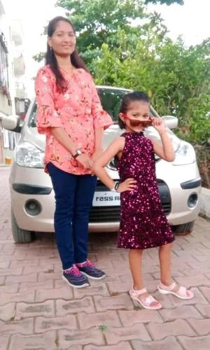Woman and young girl standing in front of a car on a paved road with trees in the background.