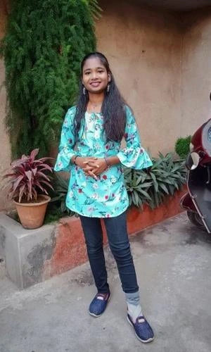 Young girl in a floral top and jeans standing outdoors with plants and a motorcycle in the background.
