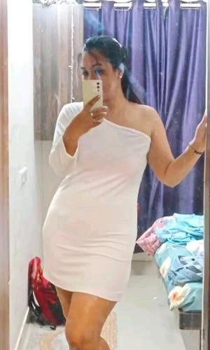 Woman taking a mirror selfie wearing a white one-shoulder dress in a room with purple curtains.