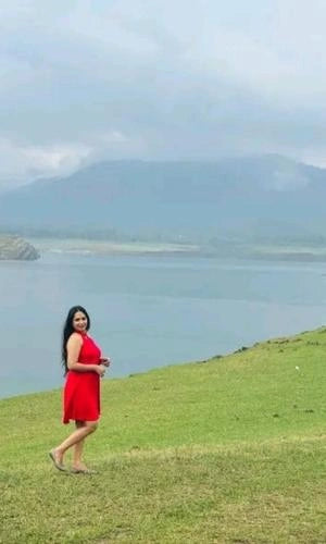 Woman in a red dress standing on a grassy hill with a lake and mountains in the background
