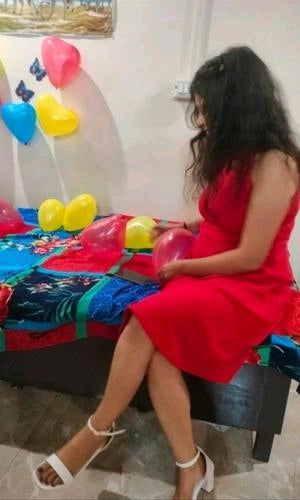 Woman in a red dress sitting on a bed with colorful balloons and a colorful quilt.