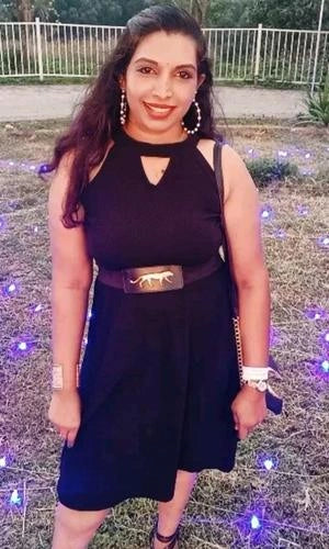 Woman in a black dress with a belt standing outdoors with purple flowers and a white fence in the background.