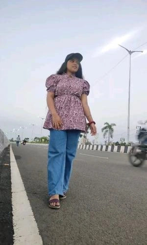 Woman walking on a road wearing a patterned top and blue pants.