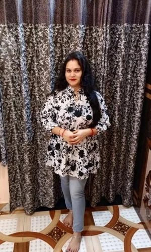 Woman wearing a black and white floral blouse with blue jeans standing in front of a patterned curtain.