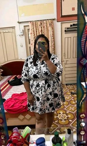 Person taking a mirror selfie wearing a black and white floral dress in a room with a bed and colorful decor.