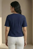 Woman wearing a navy blue top and white pants against a beige wall.