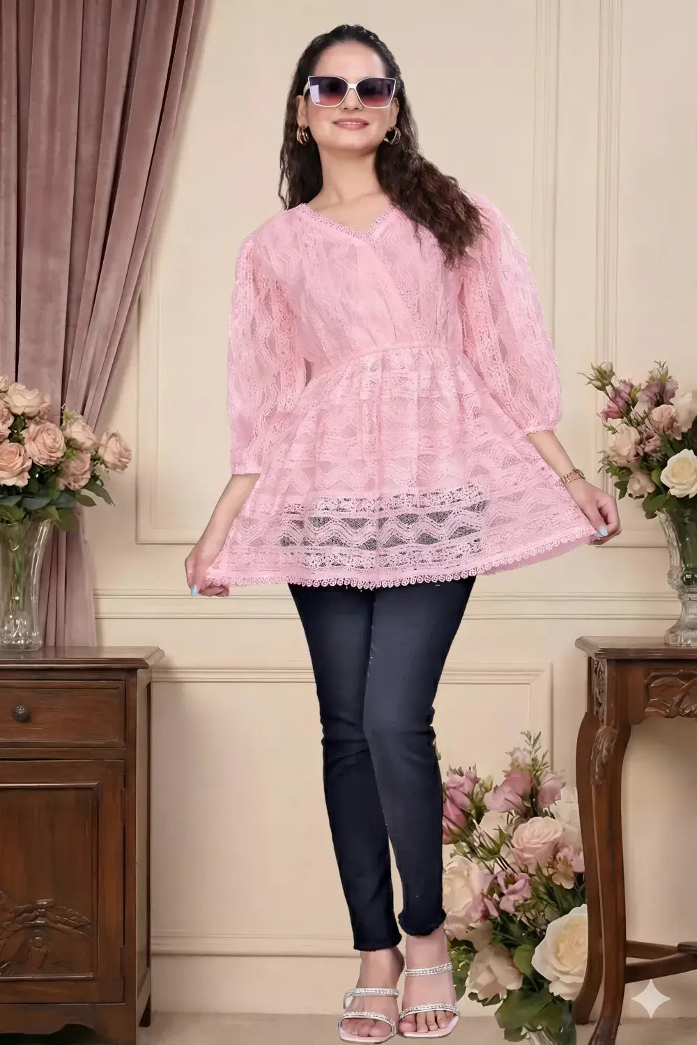 Woman wearing a pink lace top and black pants in an elegant room with flowers and furniture.
