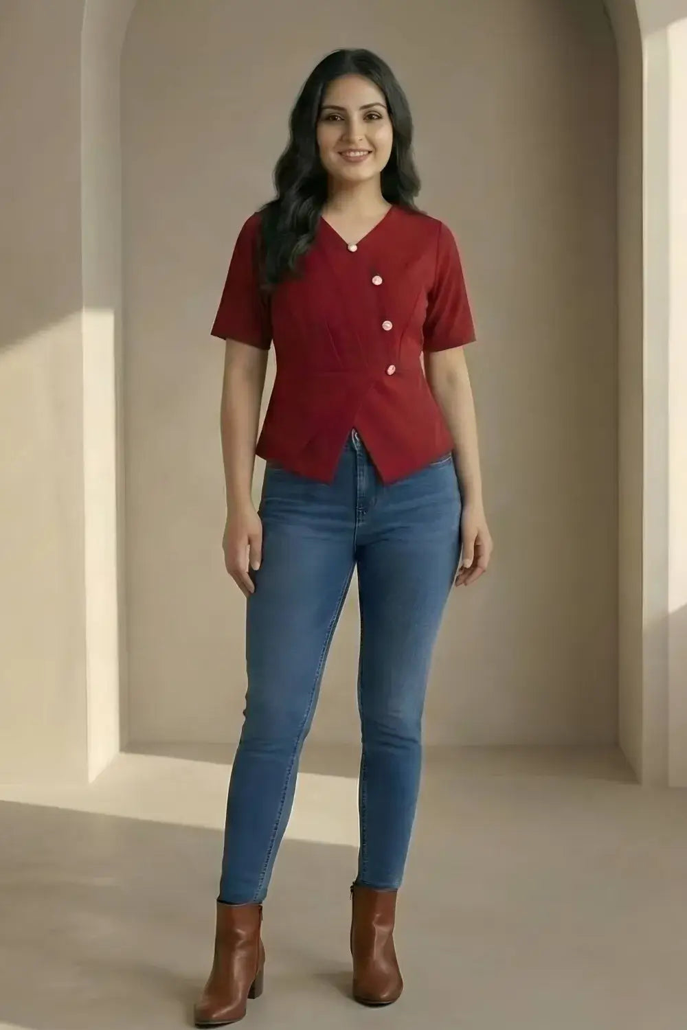 Woman wearing a red blouse with buttons and blue jeans standing in a neutral-colored room.