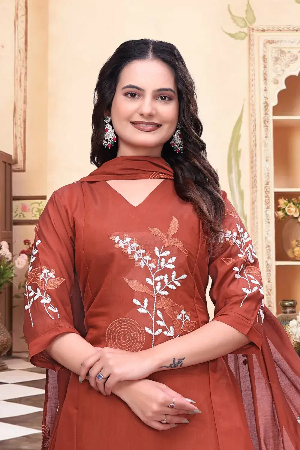 Woman wearing a rust-colored traditional outfit with floral embroidery in an indoor setting.