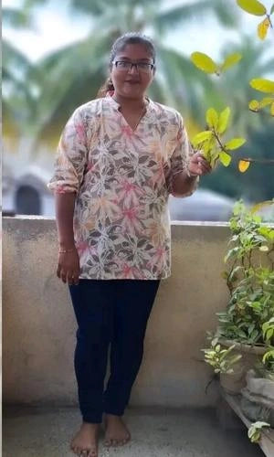 Person wearing a floral shirt and dark pants standing outdoors with greenery in the background